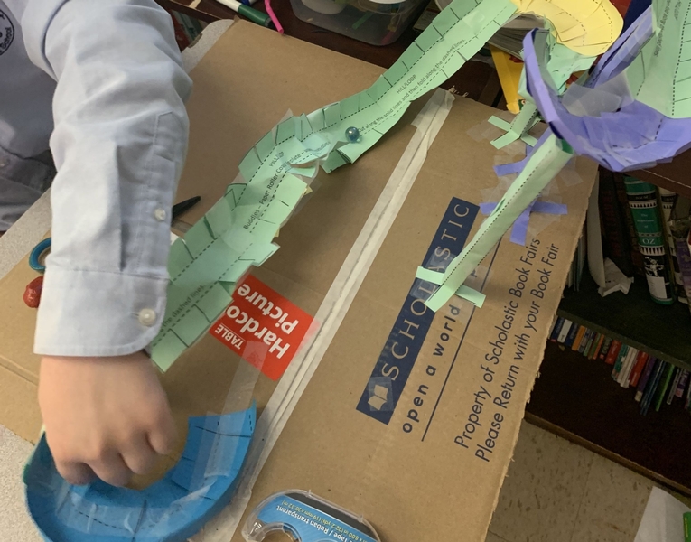 a student's arm is seen extended over box with papers cut and taped into a roller coaster shape