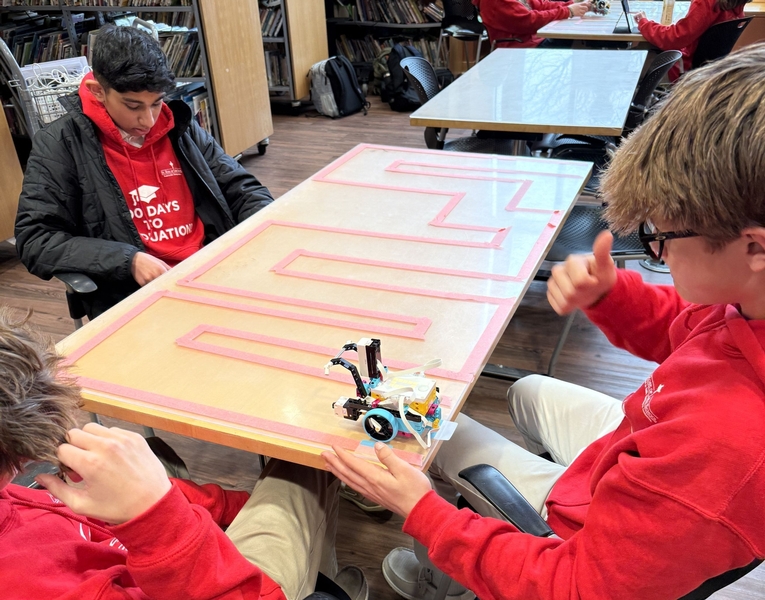 Three students sit at a table with lines of tape forming a maze. Their robot is at the start.