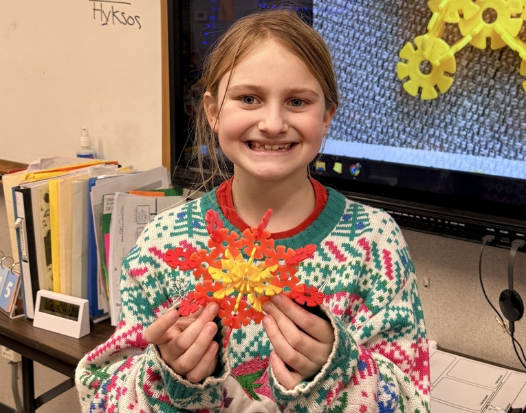 A middle grades student in a Christmas Sweater smiles holding the 3D snowflake model she built.