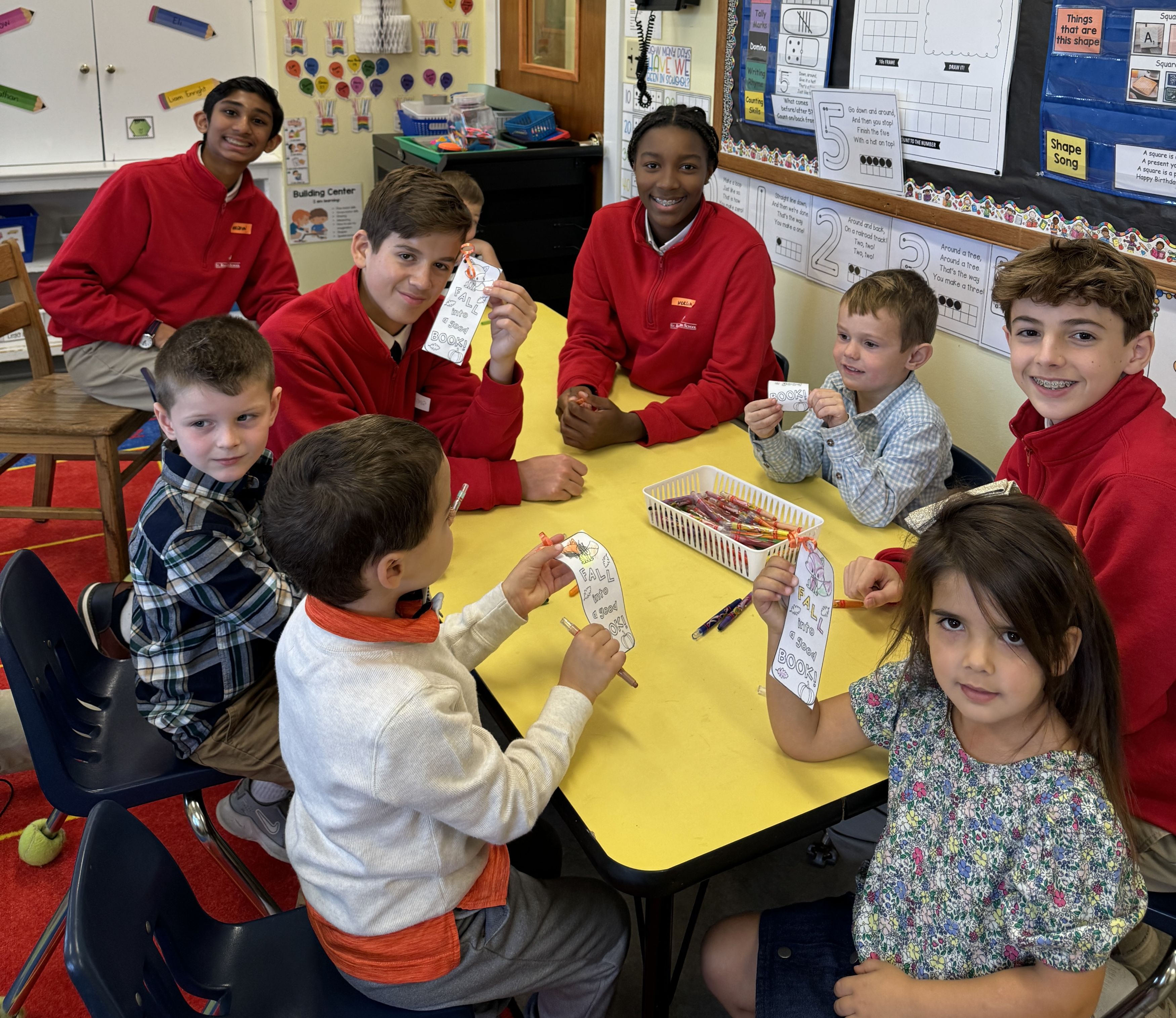Preschool students do a coloring craft around a table with their middle school buddies
